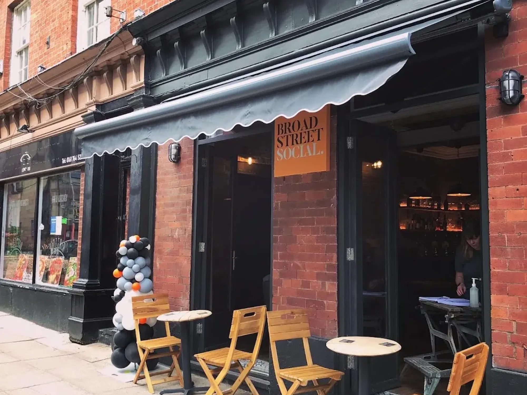 The dark painted front of Broad Street Social with its awning, orange sign and chairs on the pavement, a craft beer bar in Bury