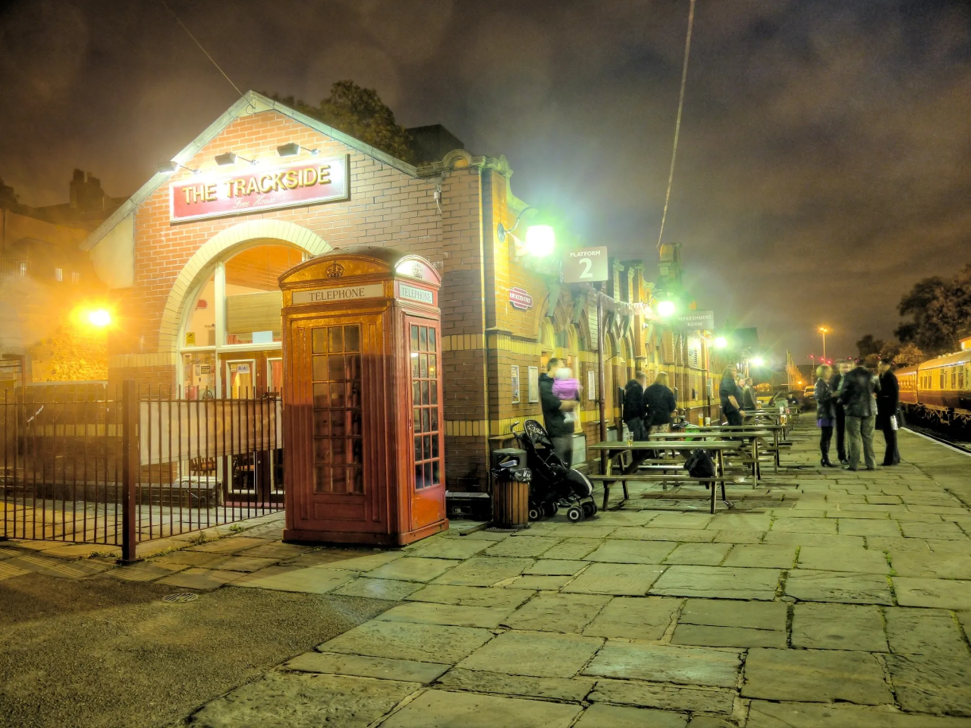 The Trackside's brick front on the platform at Bolton Street Station at night, with its sign and a red phone box, a craft beer bar in Bury