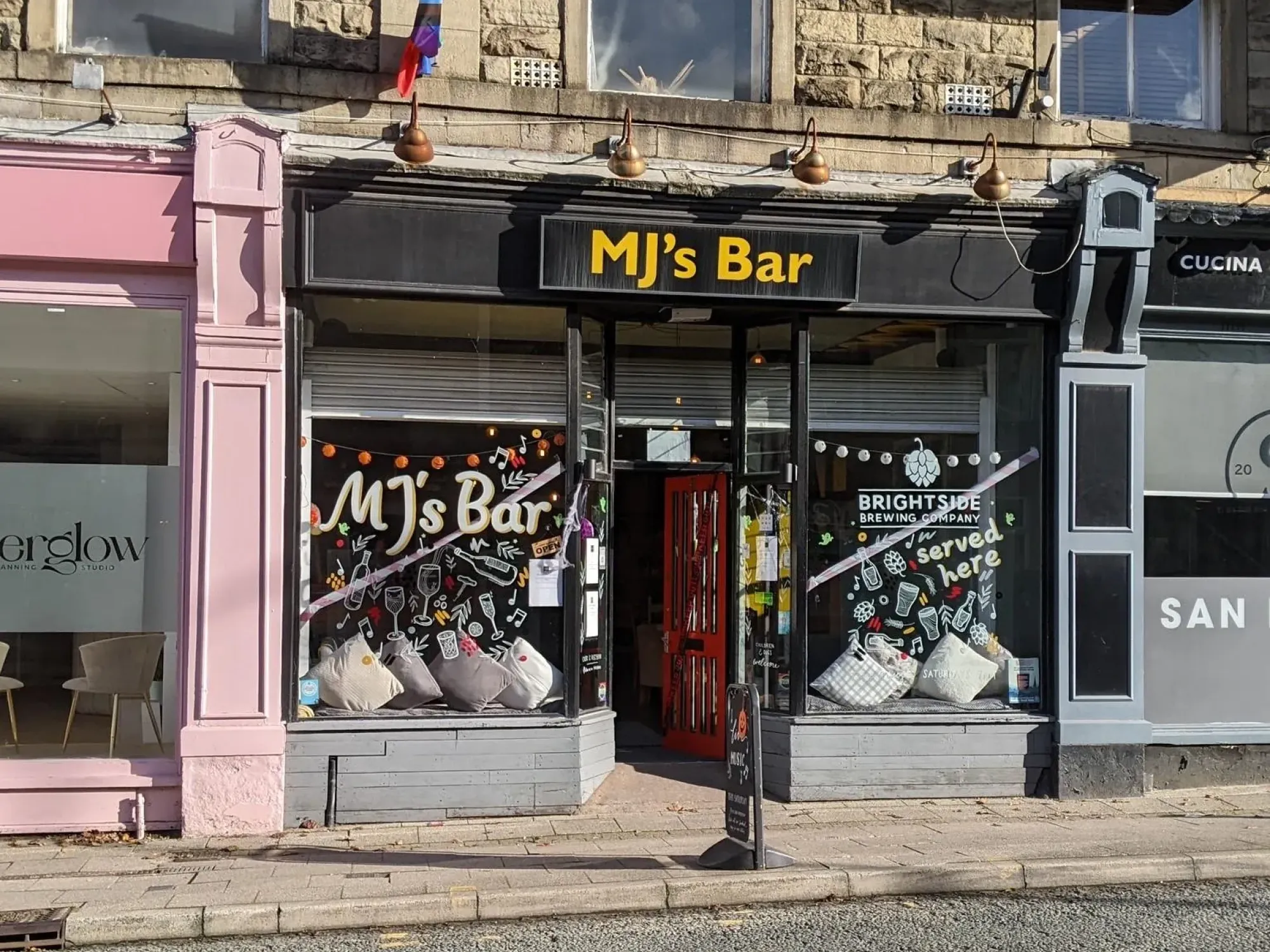 The shopfront of MJ's Bar on Bolton Street, its name over the door and painted on the window beside a red door, a craft beer bar in Ramsbottom