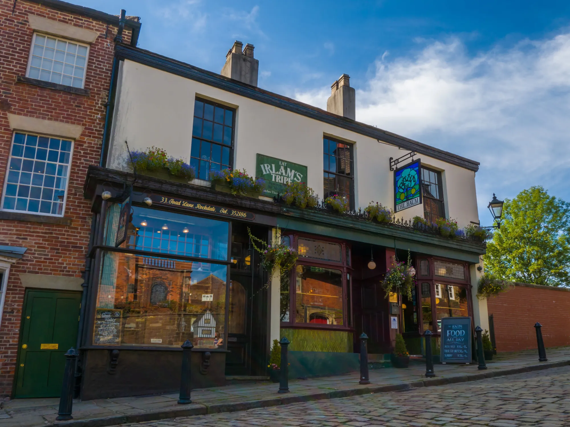 The green and cream frontage of The Baum on cobbled Toad Lane, with its hanging sign and window boxes, a pub in Rochdale