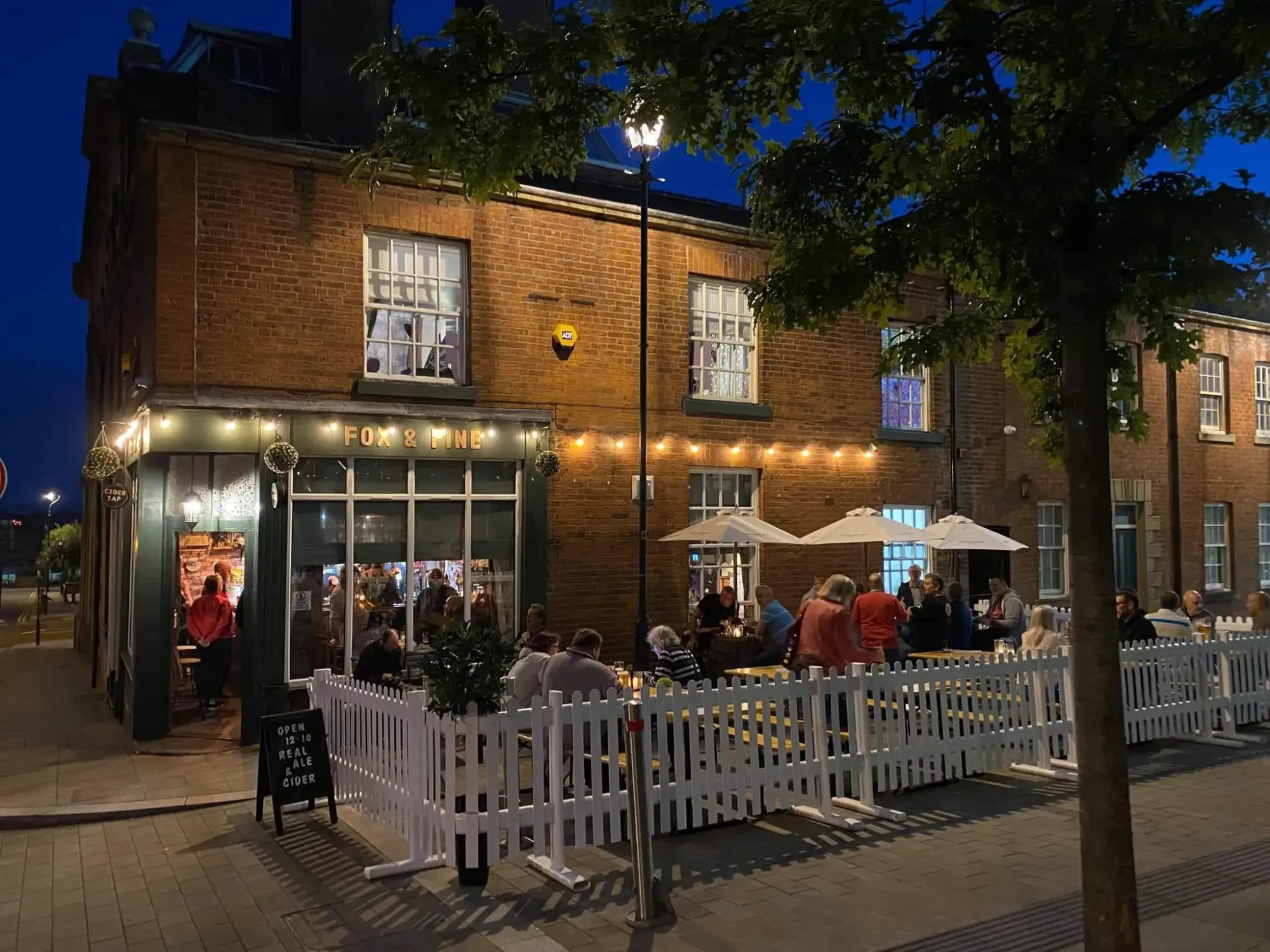 The brick corner pub at dusk with its Fox & Pine sign, festoon lights and a busy fenced terrace outside, a pub in Oldham