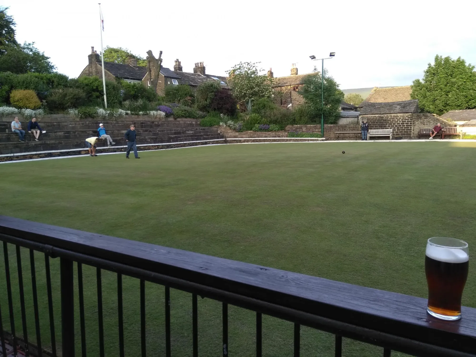 The club's bowling green on a summer evening, seen from its railed terrace with people bowling and stone cottages behind at Dobcross Band & Social Club, a pub in Dobcross