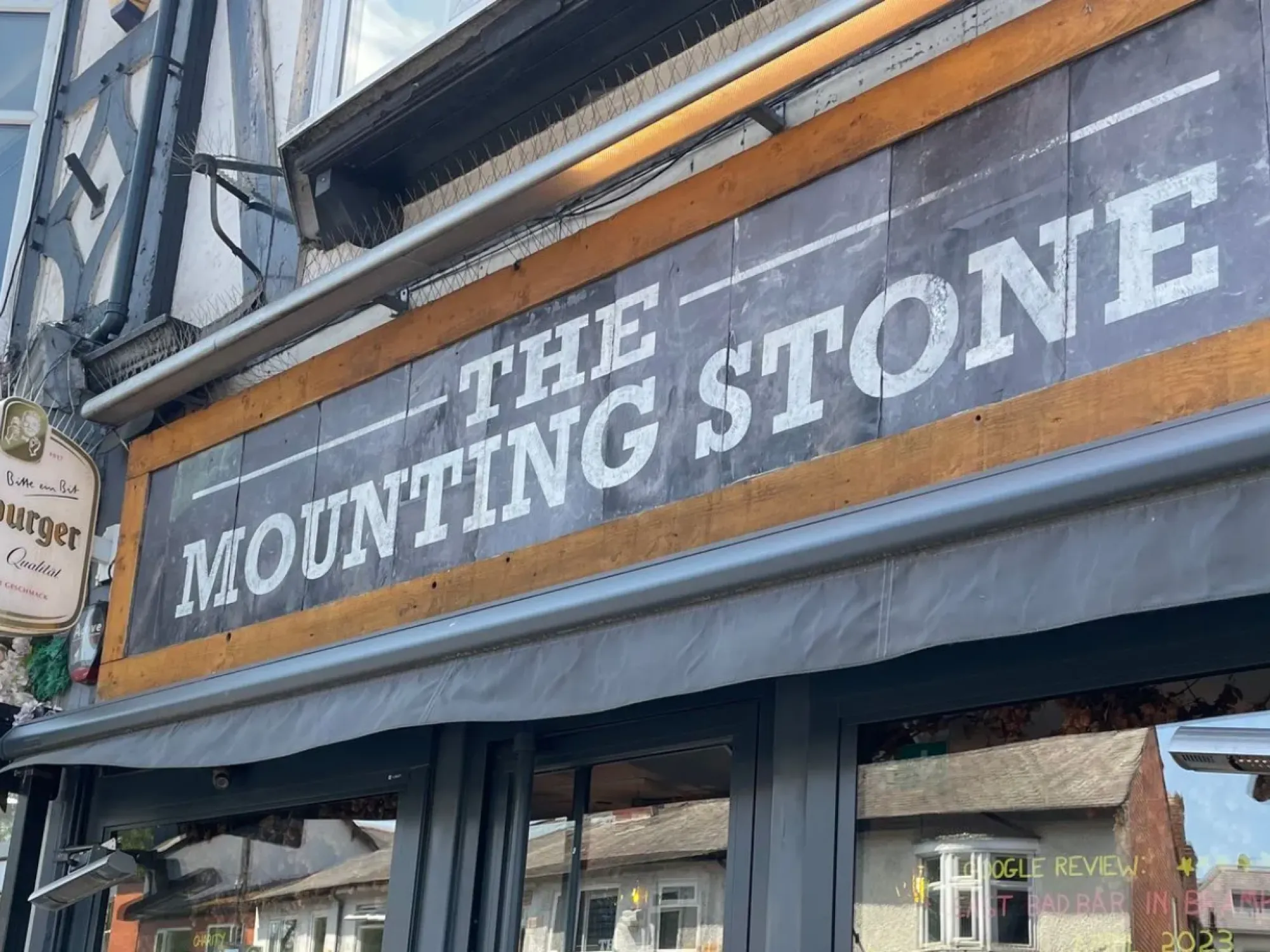 The painted Mounting Stone sign above the glazed shopfront on Woodford Road, Bramhall, a pub in Bramhall