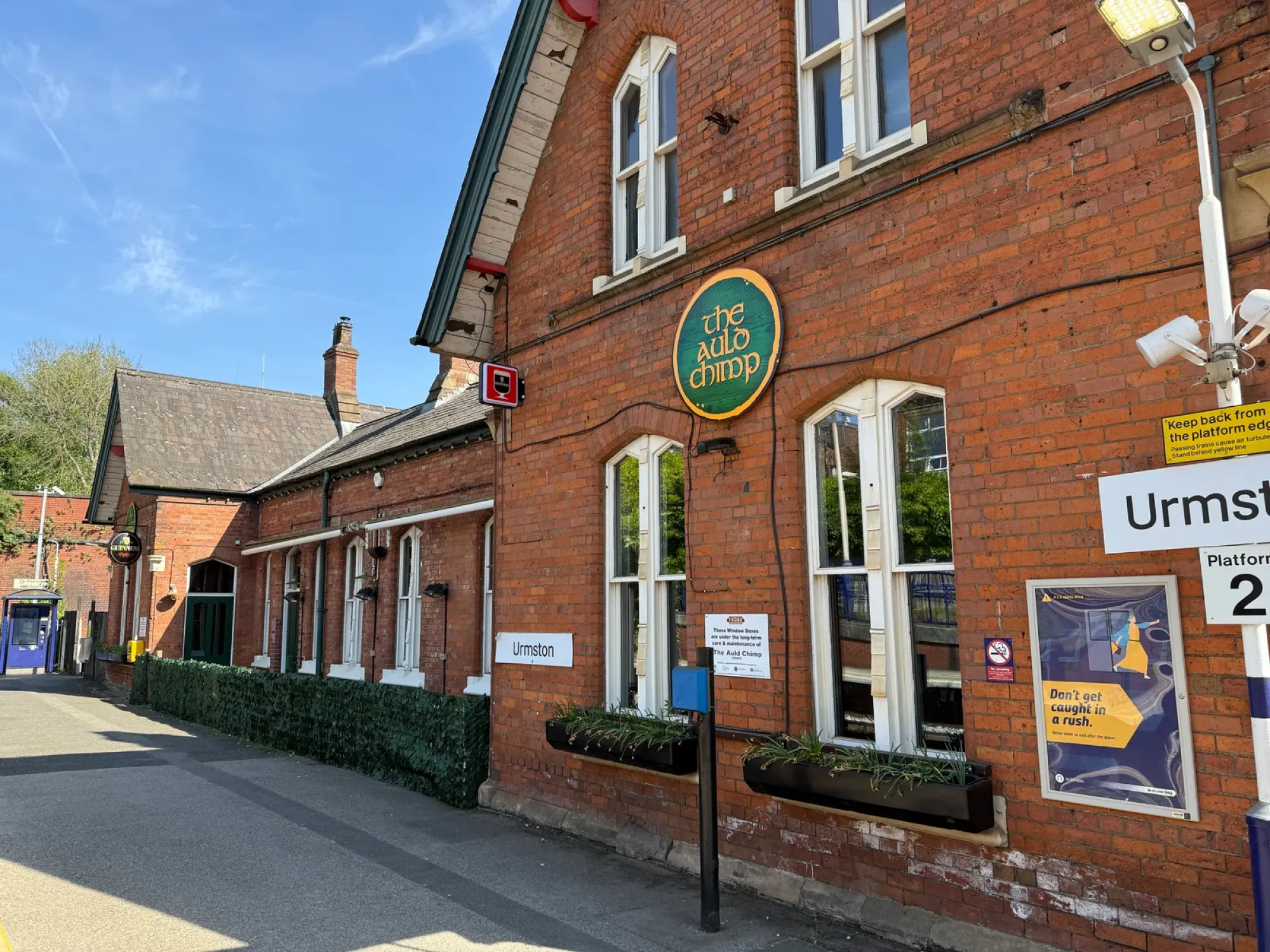 The red-brick Urmston station building with The Auld Chimp's round green sign on the wall, a pub in Urmston