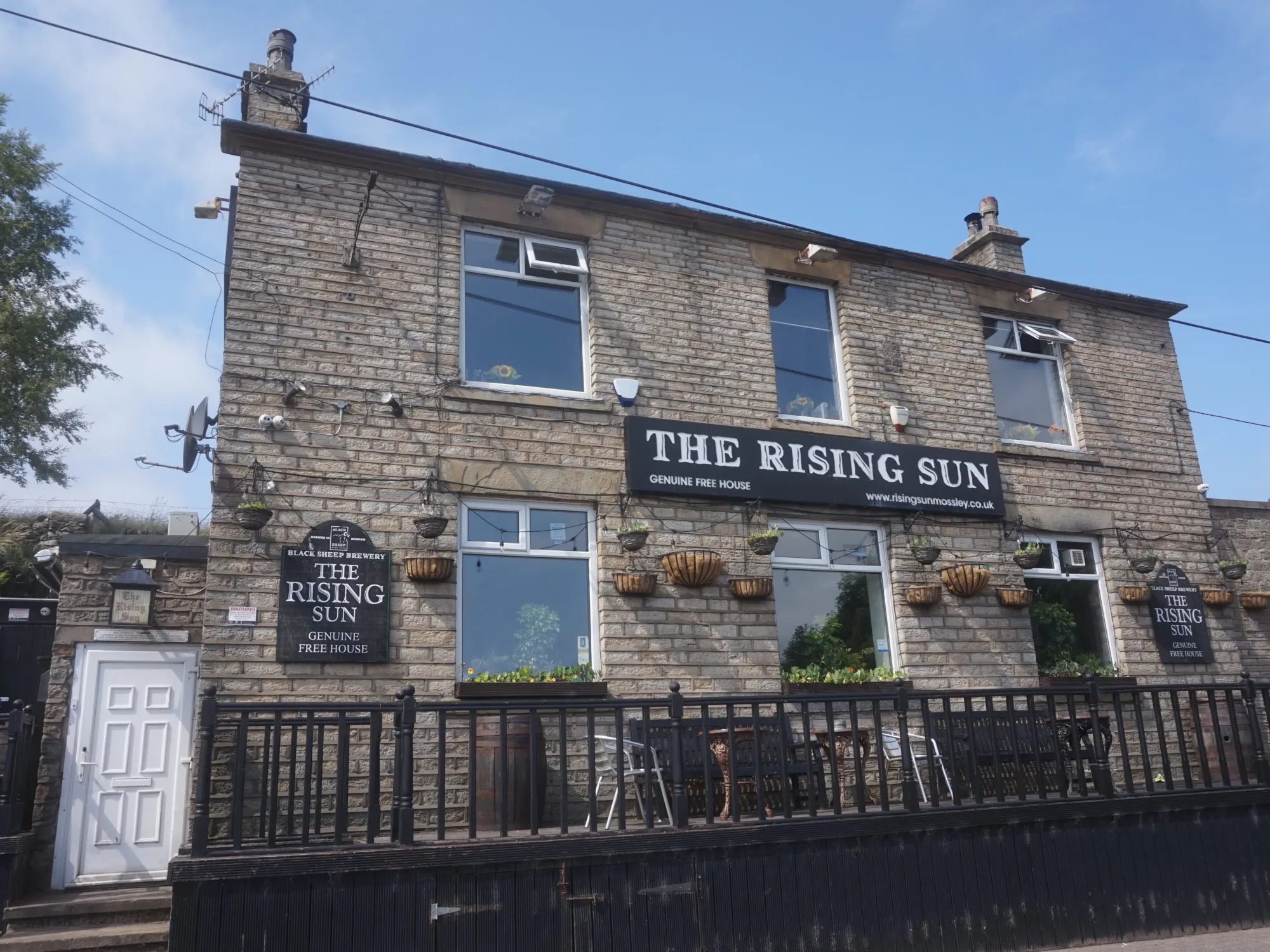 The stone front of The Rising Sun on Stockport Road, Mossley, with its sign and railed front terrace at Rising Sun, Mossley, a brew pub in Mossley