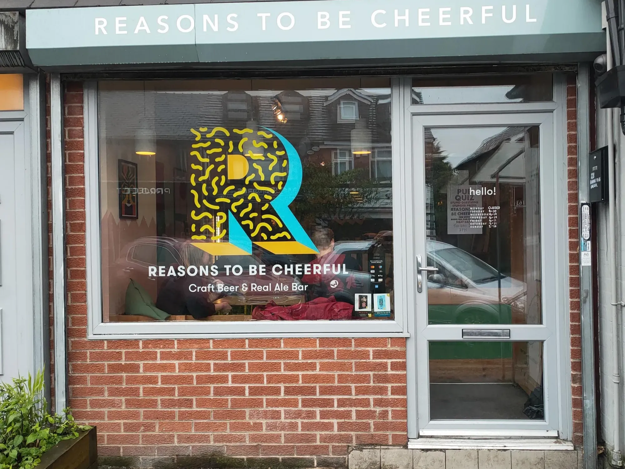 The small brick shopfront on Fog Lane, with Reasons To Be Cheerful on the fascia and a big R in the window, a craft beer bar in Burnage