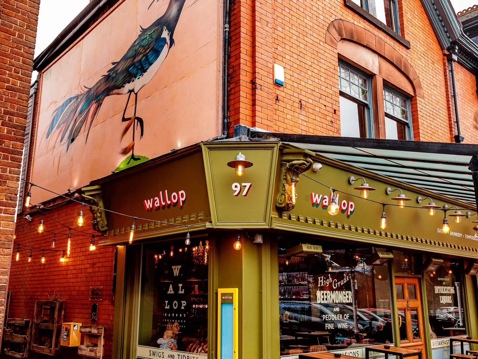 The green corner shopfront of Wallop on Lapwing Lane, with outdoor stools and a bird mural above at Wallop Didsbury, a craft beer bar in West Didsbury