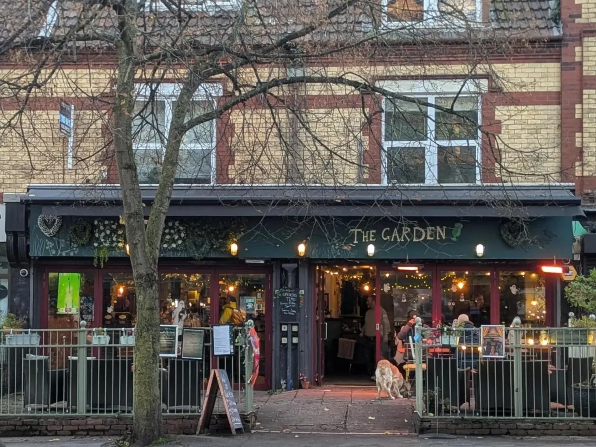 The front of the Garden on Manchester Road, lit up behind a railed terrace, with its name on the green fascia at The Garden, Chorlton, a craft beer bar in Chorlton