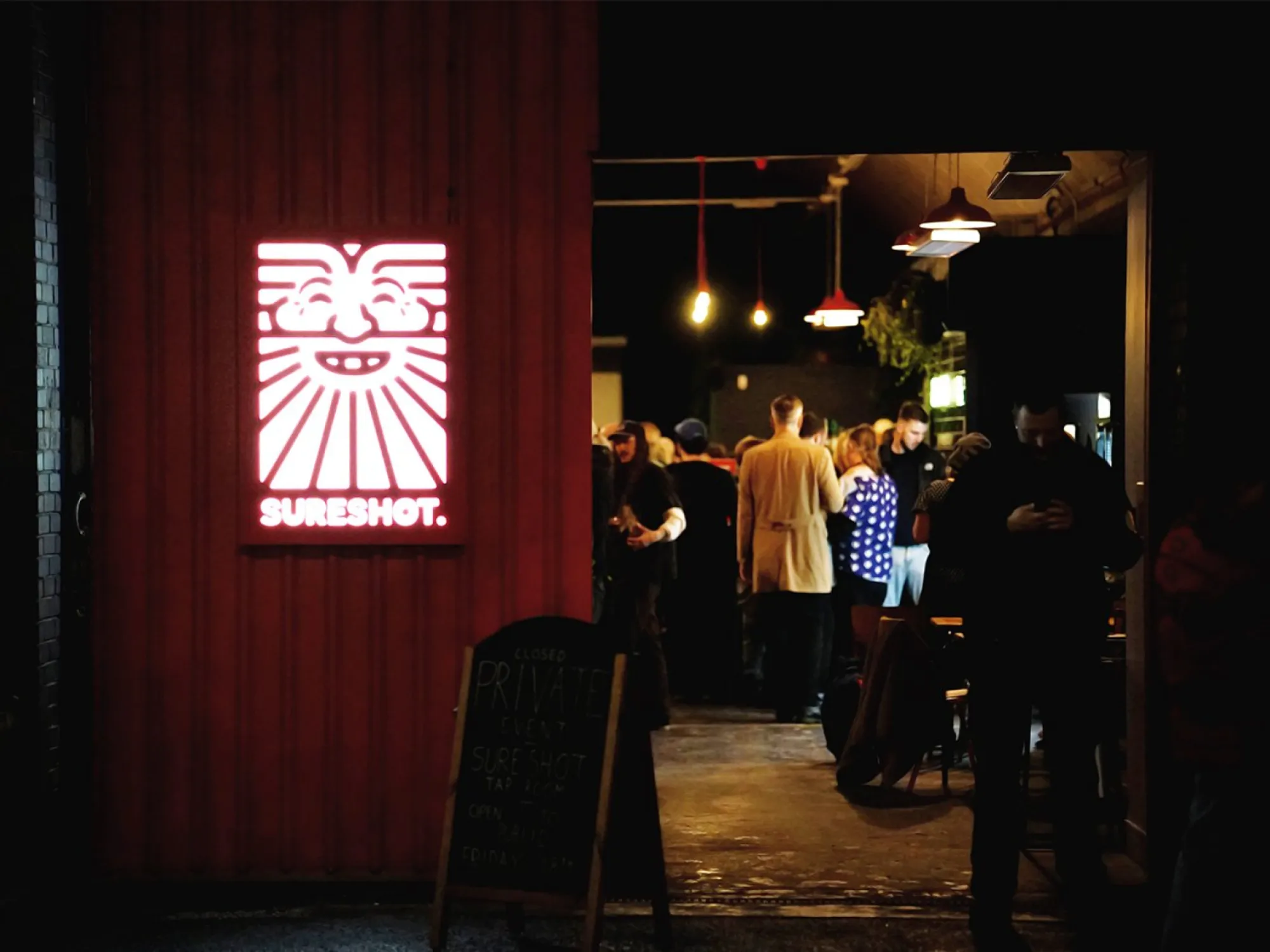 The entrance to Sureshot Taproom at night, its lit sign on the red wall and drinkers inside, a brewery taproom in Piccadilly, Manchester