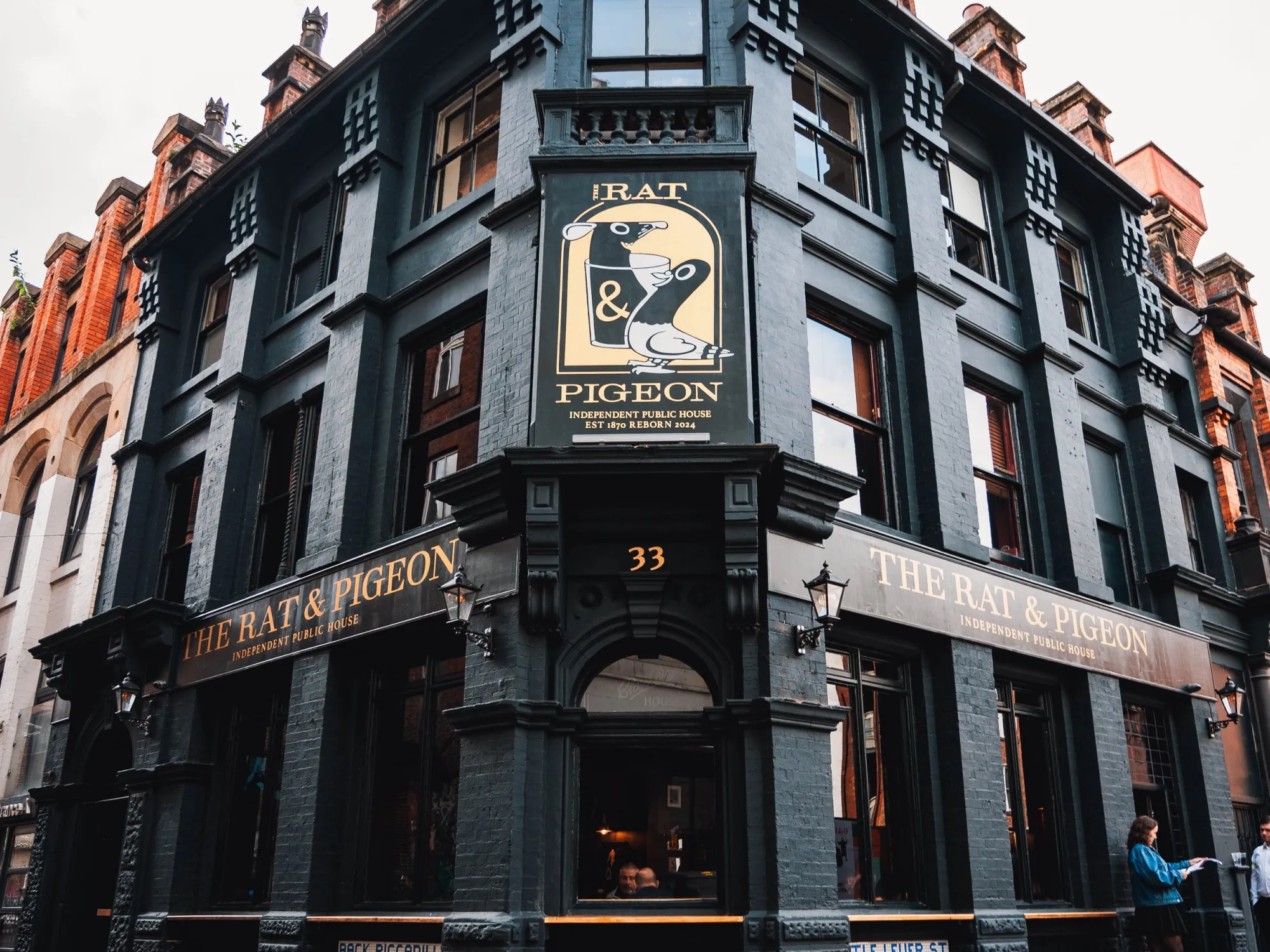 The dark painted corner front of the Rat & Pigeon on Back Piccadilly, with its gold lettered fascia and hanging sign, a pub in Piccadilly, Manchester