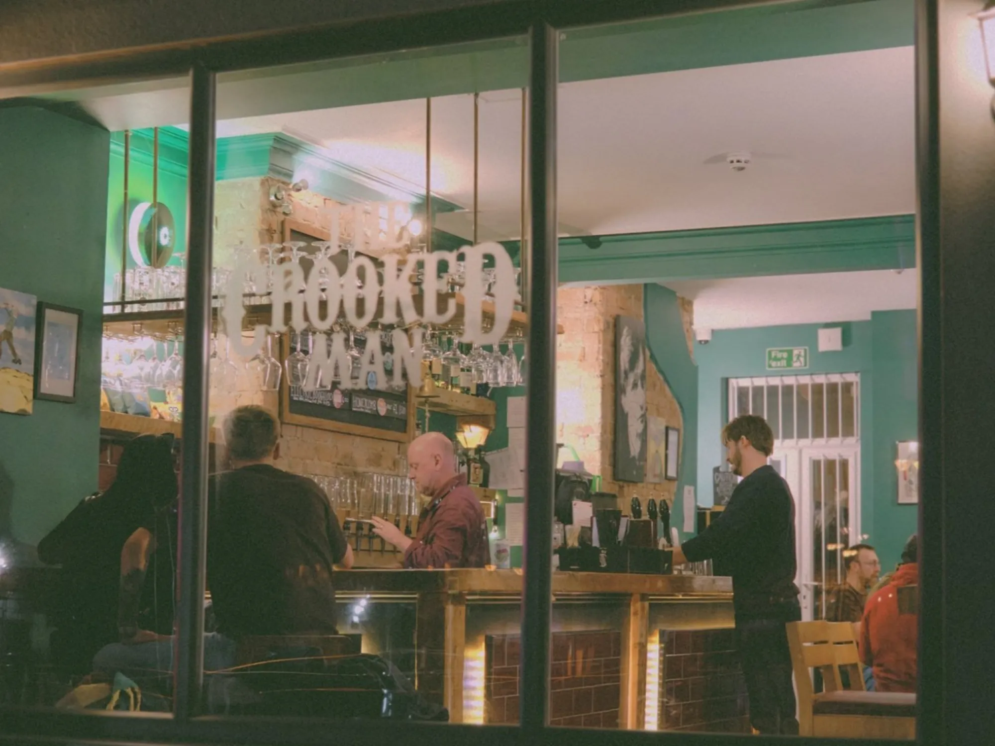 Looking through the front window of The Crooked Man at night, its name on the glass and the bar lit up inside, a craft beer bar in Prestwich, Manchester
