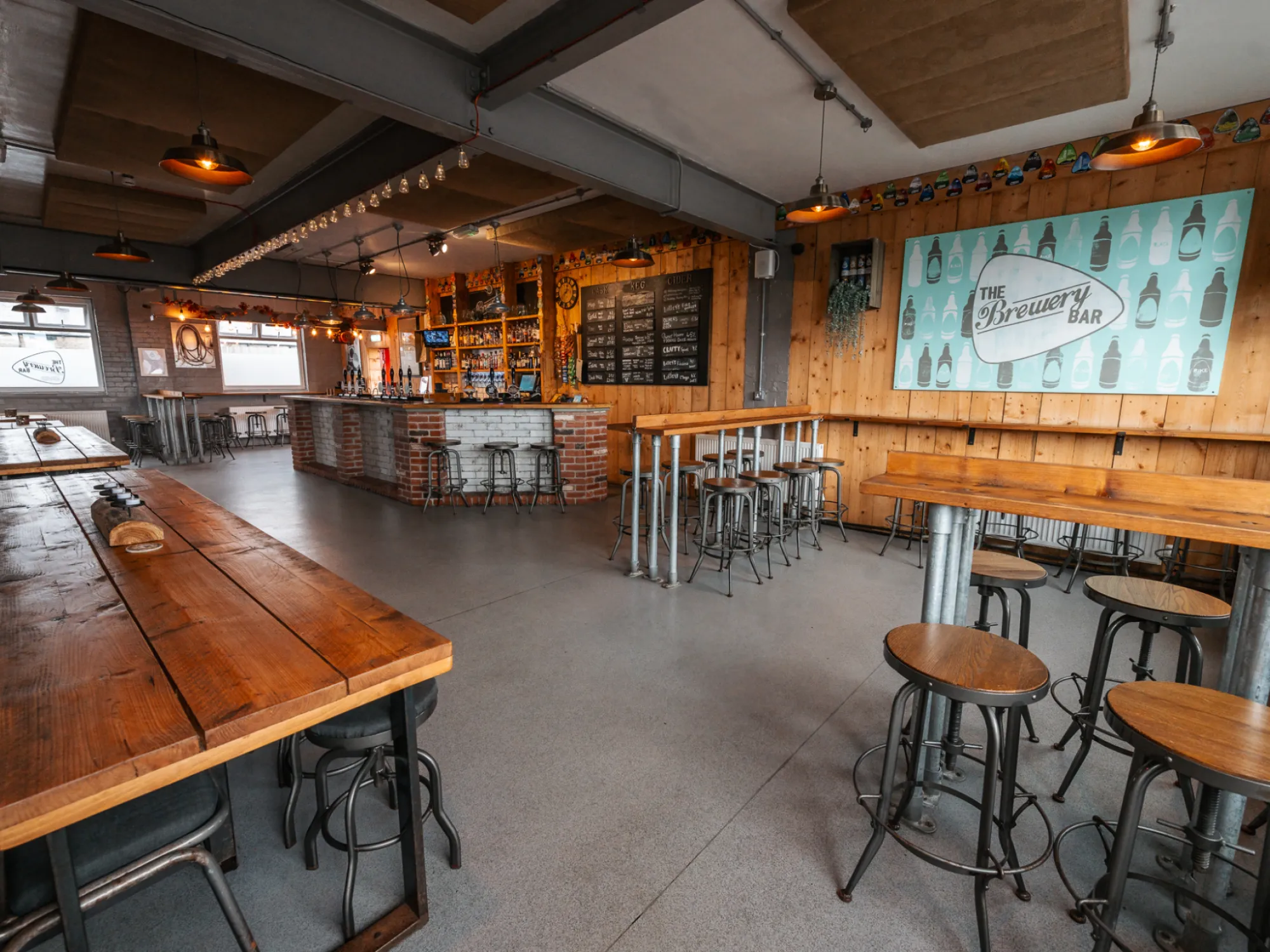 Wooden tables, stools and the brick bar inside Blackedge's Brewery Bar, with its sign on the wall, a brewery taproom in Bolton