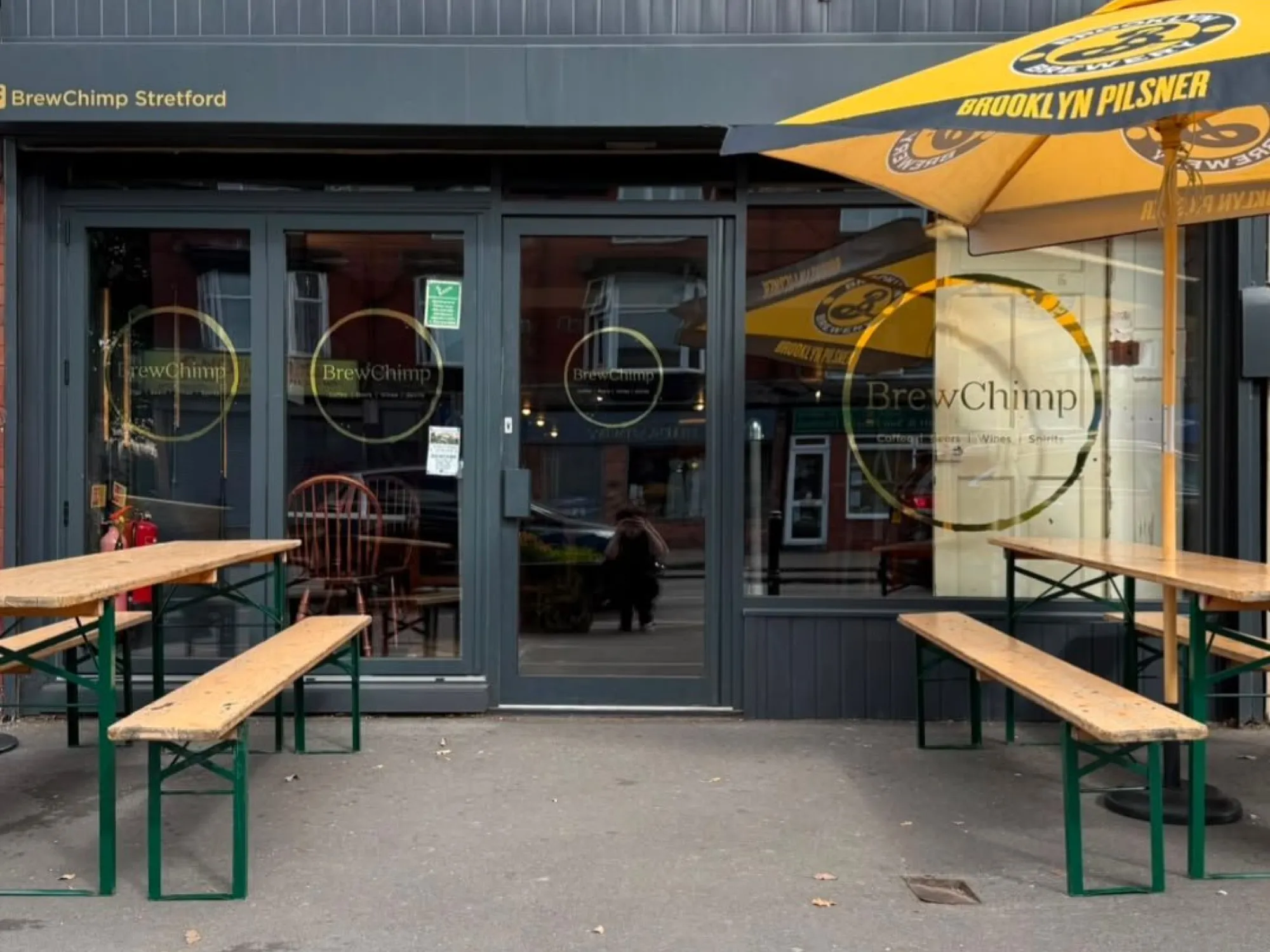 The front of BrewChimp Stretford on Barton Road, with benches and a parasol outside, a craft beer bar in Stretford, Manchester