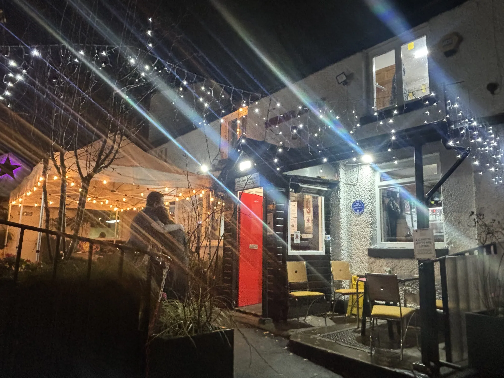 The white-painted front of the pub with its red door, blue plaque and fairy-lit courtyard at night at The Star Inn, a pub in Salford