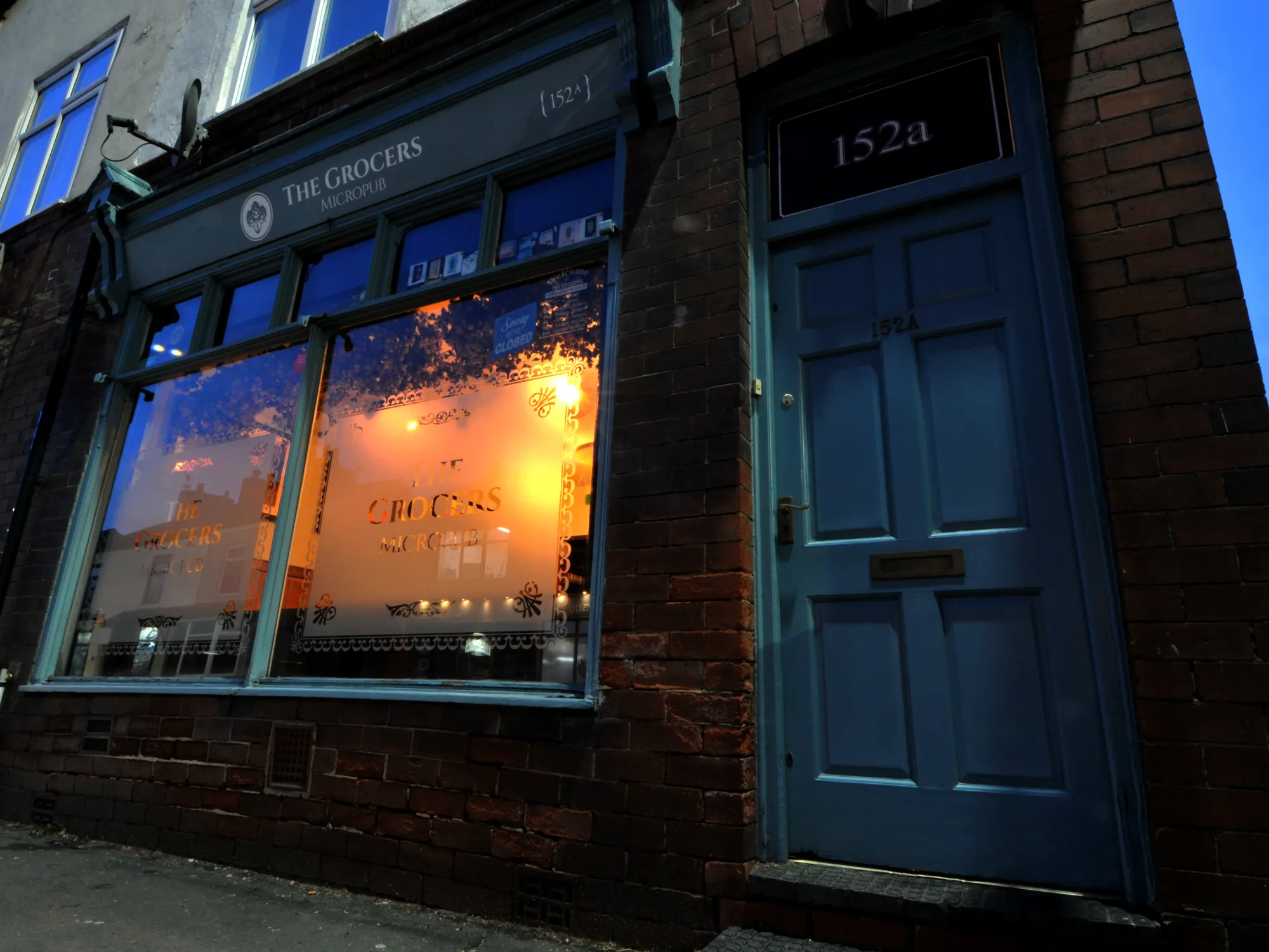 The blue-painted shopfront of the micropub at dusk, with its lit etched windows and the 152a door at The Grocers, a pub in Cadishead